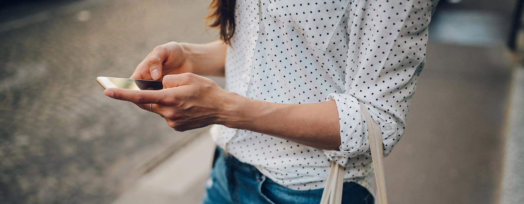 woman walks down the street and texts with a bag of groceries on her arm